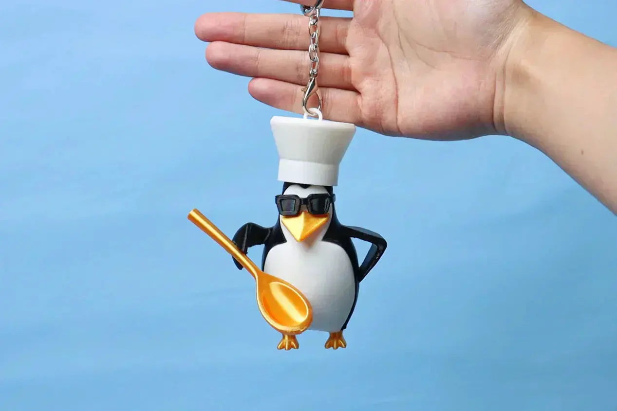 A hand holding a 3D printed keychain of a penguin wearing a chef's hat and sunglasses, holding a golden spoon