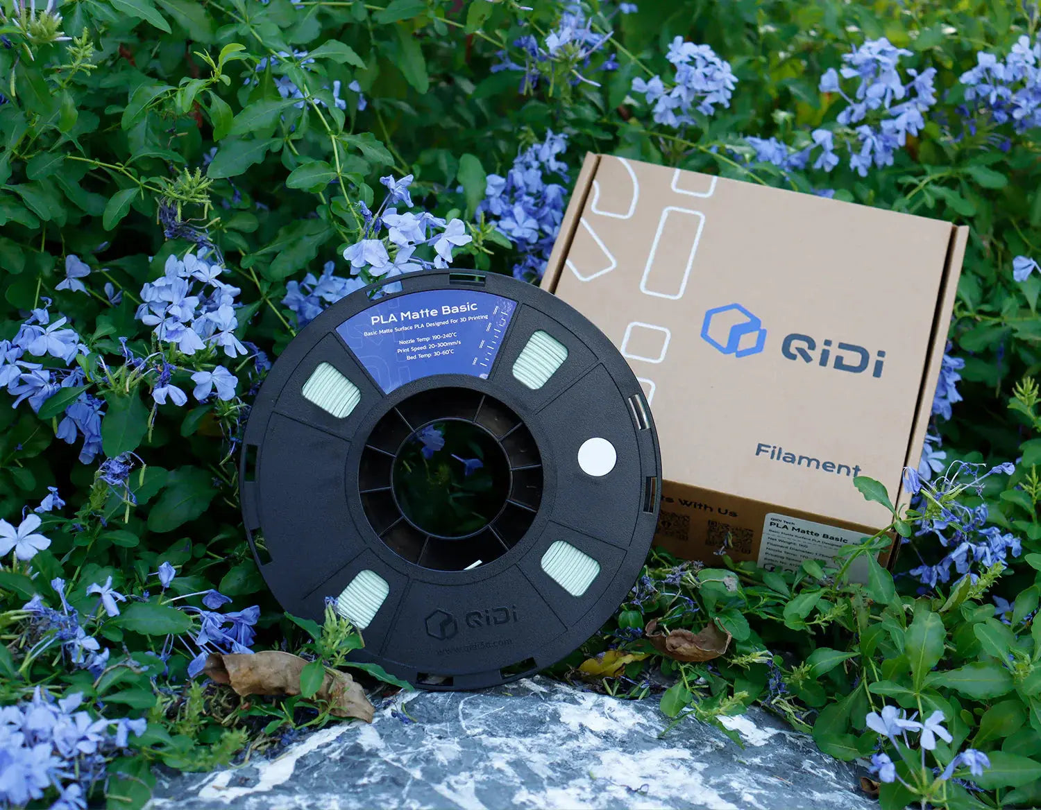 QIDI PLA  3D printer filament spool with branded box, displayed outdoors among purple flowers.