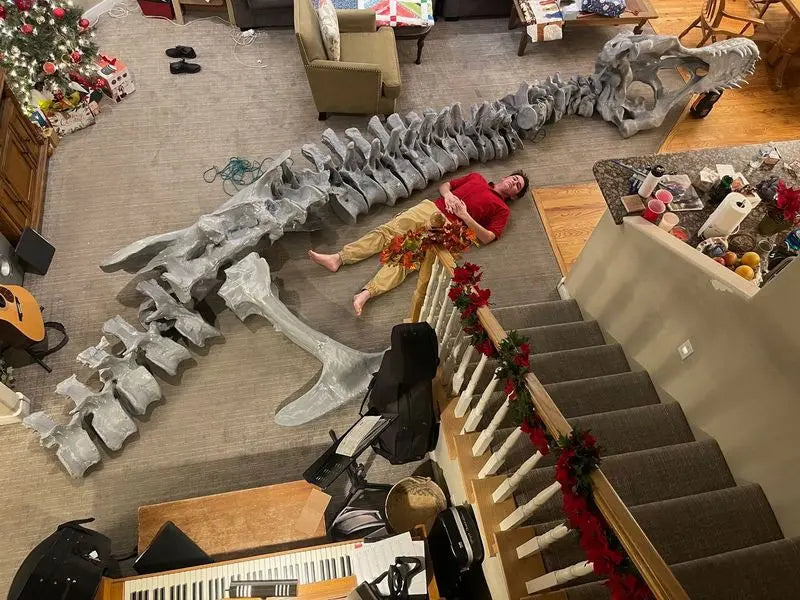 An oversized 3D printed dinosaur skeleton is spread out on the living room floor, with someone lying flat next to it for comparison, showing the overall length and volume of the model.